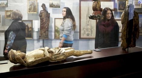 Drei Besucherinnen betrachten Kunstwerke: Holzskulpturen, darunter eine beschädigte Christusfigur, und kleinere Gemälde von Kirchen.