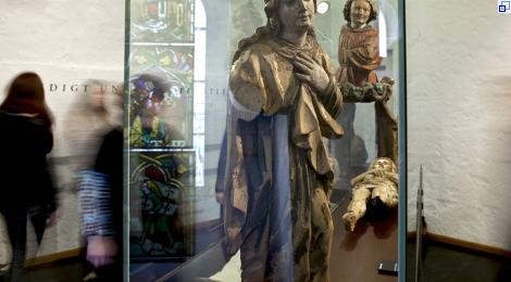Vitrine mit religiösen Holzskulpturen: eine Heilige, ein Engel und eine liegende beschädigte Christusfigur. Im Hintergrund Besucherinnen.