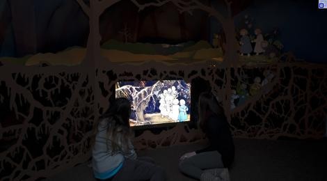 In der Wurzelhöhle im Schreiber-Museum betrachten 2 junge Frauen ein erleuchtetes Bild. Es zeigt eine Illustration aus dem Kinderbuch "Etwas von den Wurzelkindern". Das Motiv ist die Wurzelhöhle unter der Erde: Unter dicken Wurzeln laufen die Wurzelkinder in die Höhle hinein.