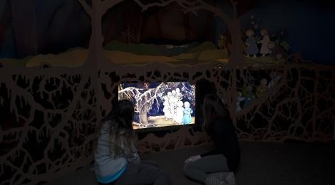 In der Wurzelhöhle im Schreiber-Museum betrachten 2 junge Frauen ein erleuchtetes Bild. Es zeigt eine Illustration aus dem Kinderbuch "Etwas von den Wurzelkindern". Das Motiv ist die Wurzelhöhle unter der Erde: Unter dicken Wurzeln laufen die Wurzelkinder in die Höhle hinein.