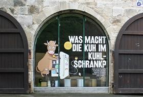 Großes Fenster mit dem aufgeklebten Plakatmotiv der Ausstellung, einer gezeichneten Kuh vor einem Kühlschrank.