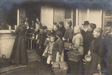 Historisches Schwarzweiß-Foto: Erwachsene und Kinder mit Gefäßen stehen Schlange vor einen offenem Fenster und einer geöffneten Tür.