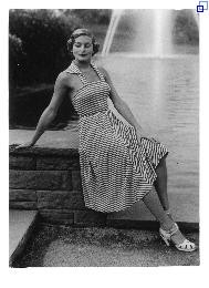 Eine Frau posiert auf einer kleine Mauer sitzend in einem Sommerkleid. Im Hintergrund eine Wasserfläche mit Fontäne. 