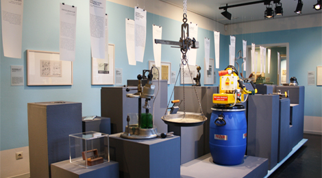 Ausstellungsraum mit vielen Sockeln, auf denen vor allem technische Geräte stehen. Über den Gegenständen hängen (wie in der ganzen Ausstellung) Blätter mit Informationen zu den einzelnen Objekten. 