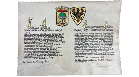 Eine prächtig gestaltete Urkunde. Sie zeigt zwei Wappen, darunter handgeschriebener Text in zwei Blöcken, links französisch, rechts deutsch. Unter dem Text die Unterschrift der beiden Bürgermeister.