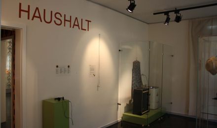 Ausstellungsbereich "Haushalt", unter anderem mit einer Kittelschürze und einer Schleuder sowie mit einer Hörstation.