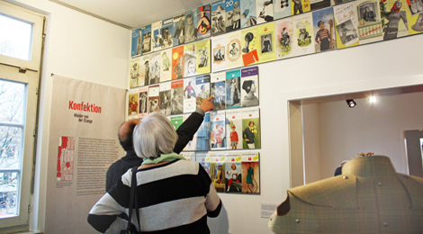 Ein Besucher zeigt einer Besucherin etwas auf einer Wand, an der viele Anleitungshefte der "Esslinger Wolle" aufgehängt sind.