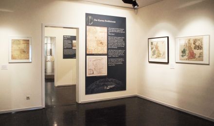 Ausstellungsraum mit gerahmten Karten an der Wand und großer Texttafel "Der Kartenperfektionist".