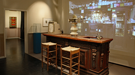 Ausstellungsraum: Wirtshaustresen mit prunkvoller Zapfanlage, davor zwei Stühle, dahinter eine Projektion einer typischen Kneipenwand mit Flaschen.