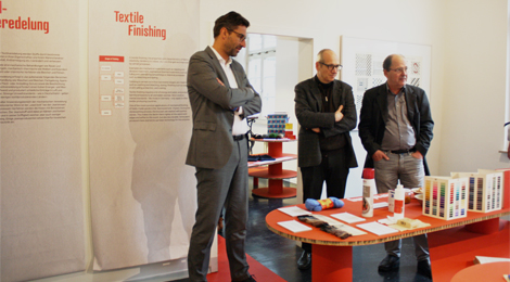Drei Männer betrachten Ausstellungsobjekte auf einem Podest.