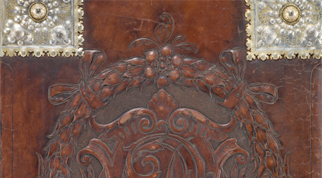Foto: Michael Saile (Ausschnitt). Detailfoto einer Ledermappe mit Teil eines geprägten Emblems, daneben verzierte silberne Metallbeschläge .