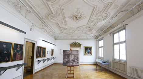 Patrizierzimmer mit Stuckdecke und Gemälden an den Wänden.