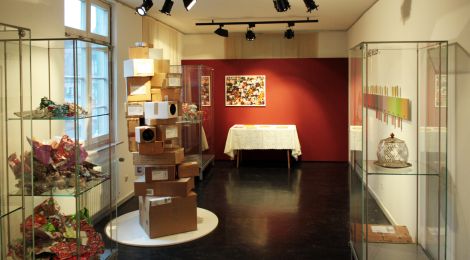 Ausstellungsraum mit einer Installation aus übereinander geschachtelten Versandkartons. Daneben steht eine Vitrine mit geknülltem Geschenkpapier.