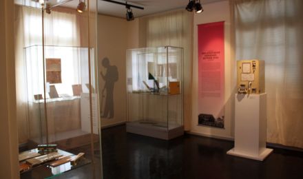 Ausstellungsraum mit Vitrinen und einer großen Texttafel. Im Mittelpunkt steht ein alter Fahrkartenautomat mit Handkurbel.