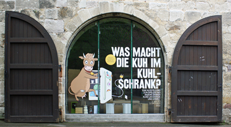 Großes Fenster mit dem aufgeklebten Plakatmotiv der Ausstellung, einer gezeichneten Kuh vor einem Kühlschrank.