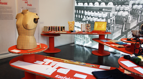 Ausstellung "Garne, Stoffe, Waren": Ausstellungsraum mit Objekten auf Podesten und einem großen Bild eines Fabrikraumes im Hintergrund.