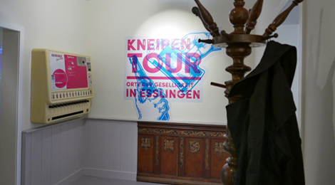 Ausstellungsraum: Ein Garderobeständer mit Mantel und ein Zigarettenautomat, dahinter das Ausstellungsplakat. und 