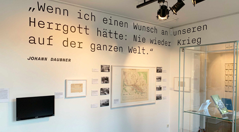 Ausstellungsraum: An der Wand hängen eine Landkarte und mehrere Fotos. Auf der Wand steht groß das Zitat: "Wenn ich einen Wunsch an unseren Herrgott hätte: Nie wieder Krieg auf der ganzen Welt."