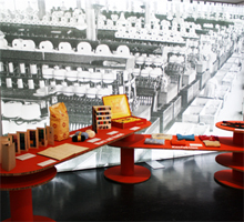 Ausstellungsraum mit Objekten auf Podesten und einem großen Bild eines Fabrikraumes im Hintergrund.