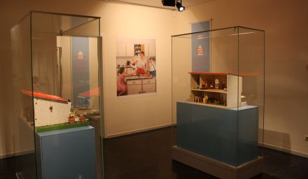 Ausstellungsraum: Vitrinen mit Puppenhäusern im Stil von modernen Häusern der 1960er Jahre. An der Wand hängt die vergrößerte Reproduktion einer Anzeige für Küchengeräte.