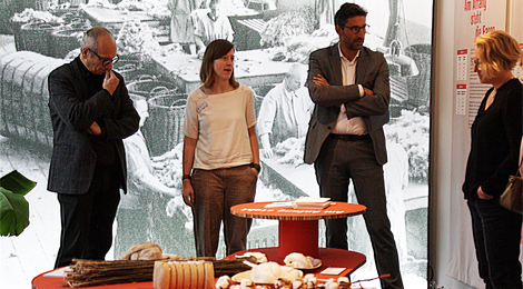 Vier Personen stehen vor einem Podest mit Ausstellungsobjekten. Im Hintergrund ein ein wandgroßes beleuchtetes Foto von einer Fabrikhalle.