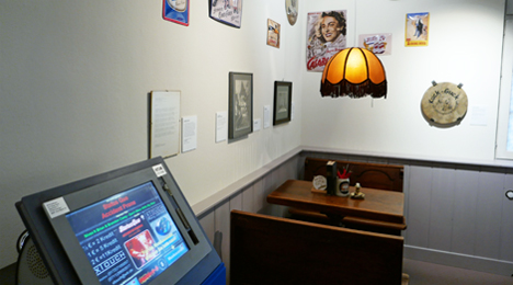 Ausstellungsraum: Ein Tisch mit zwei Bänken und Deckenlampe, davor der Computerbildschirm einer Jukebox. An der Wand hängen  Filmplakate, Bilder und Reklameschilder.