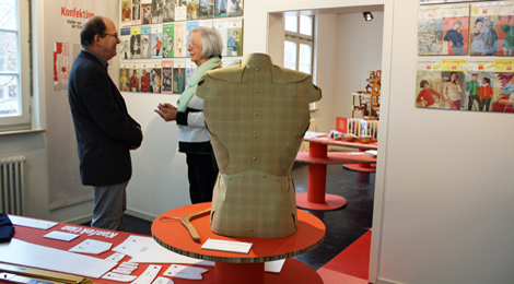 Zwei Personen stehen zwischen einem Podest mit Ausstellungsobjekten und an der Wand aufgehängten Heften mit Strickanleitungen. Sie unterhalten sihc.