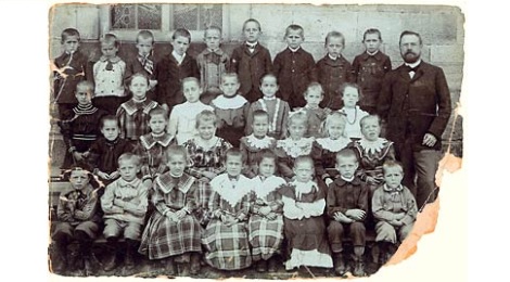 Ein sehr altes schwarzweißes Klassenfoto mit Lehrer, Mädchen und Jungen. Das Foto ist beschädigt, eine Ecke fehlt.