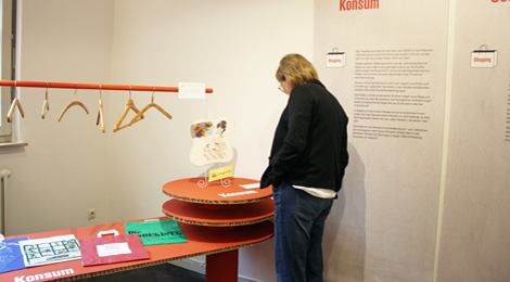 Eine Besucherin liest eine Objektbeschriftung. Neben ihr an der Wand ein Text mit der Überschrift "Konsum".