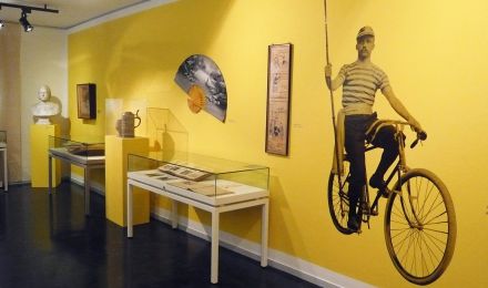 Ausstellungsraum mit Vitrinen und als Schmuck ein aufgeklebtes lebensgroßes Bild eines Radfahrers an der Wand.
