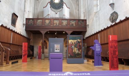 Chor der Franziskanerkirche als Ausstellungsraum. Blick nach hinten zur Orgelempore. Im Raum verteilt stehen eine Stellwand mit einem großen Gemälde und mehrere Schrift-Tafeln.