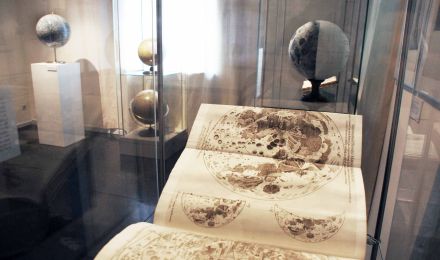 Ausstellungsraum: In einer Vitrine liegt ein großes aufgeschlagenes Buch mit ausgeklappten Karten, die den Mond abbilden.Im Hintergrund der Mondglobus von Tobias Mayer.