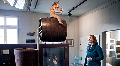 Foto: Daniela Wolf (Ausschnitt). Stadtmuseum: Eine Besucherin betrachtet ein Weinfass, auf dem rittlings eine Bacchus-Skulptur sitzt.