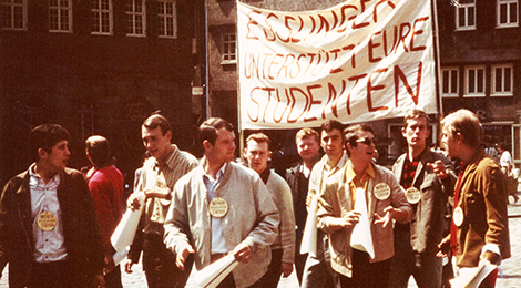 Foto aus den 1960er oder 1970er Jahren von einer Studentendemonstration: Junge Männer gehen in einer Gruppe, sie halten ein beschriftetes Transparent hoch.